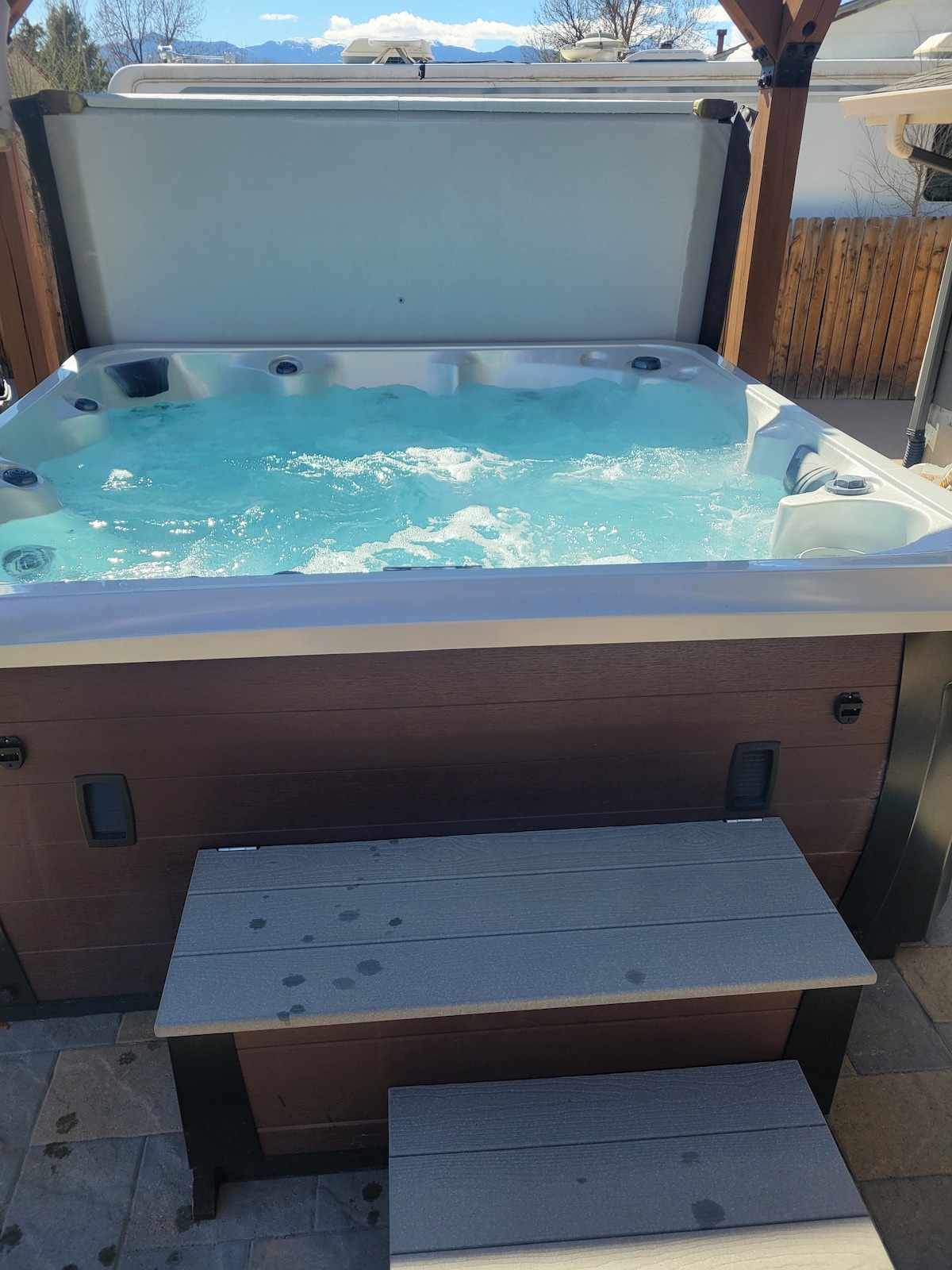A hot tub is positioned under a wooden pergola, featuring a spacious seating area along one side. Water bubbles gently in the tub, inviting relaxation. Surrounding natural elements are visible in the backdrop, contributing to a serene outdoor atmosphere.