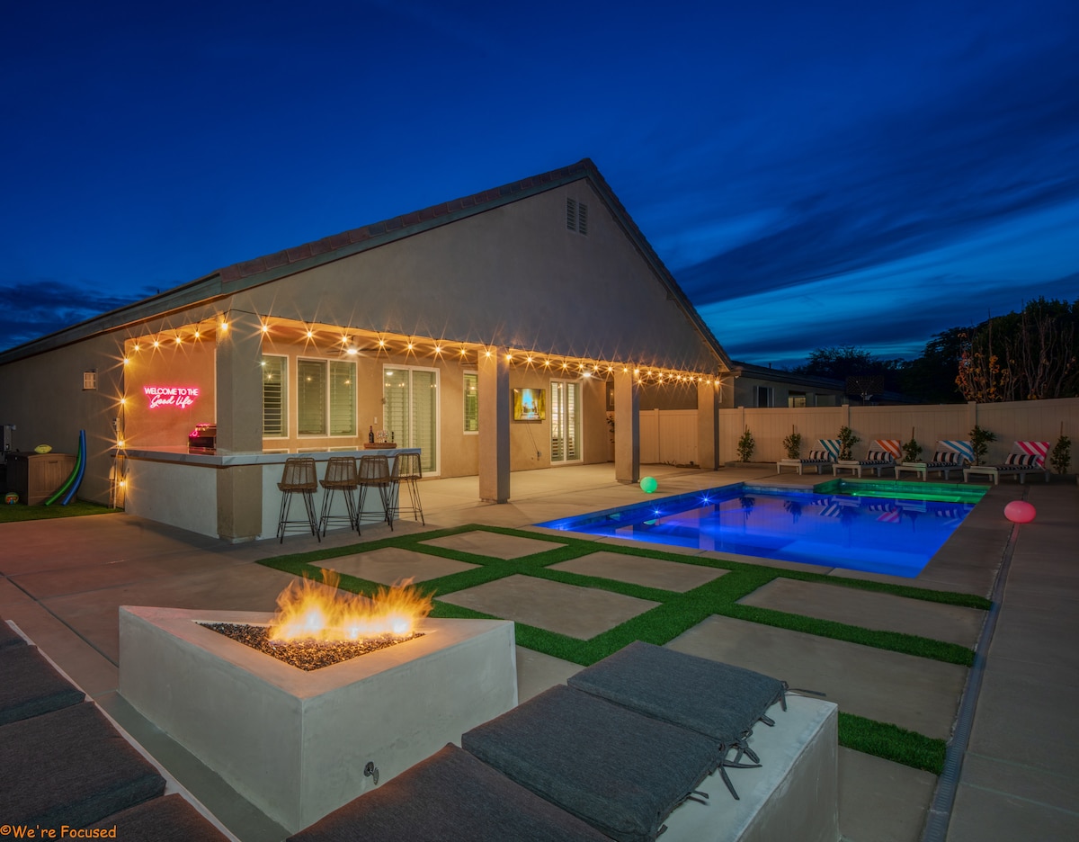 An outdoor space is illuminated by string lights, featuring a rectangular pool surrounded by lounge chairs. A fire table is centered on the patio, providing warmth. The house, visible in the background, showcases large windows reflecting the evening sky.