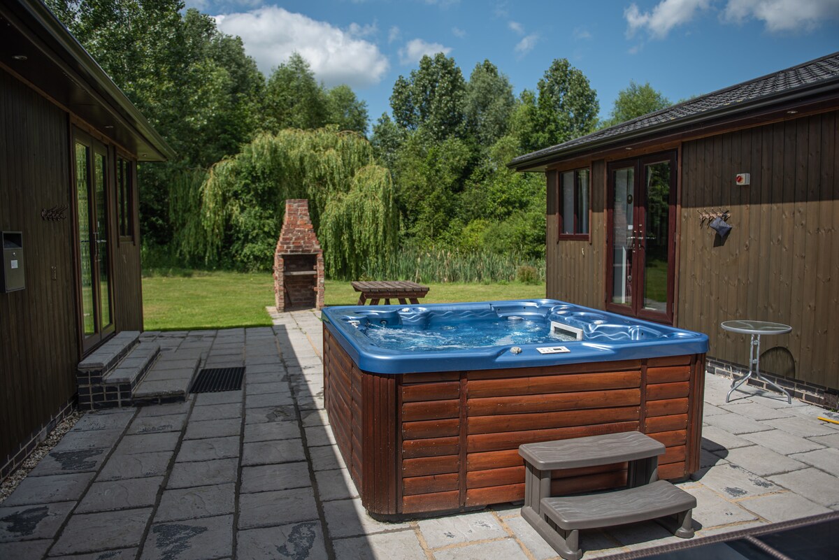 A hot tub is surrounded by a spacious stone patio, bordered by grass and trees. A brick fireplace stands nearby, and a picnic table is positioned for outdoor dining. Sunlight filters through the clouds, enhancing the serene outdoor setting.