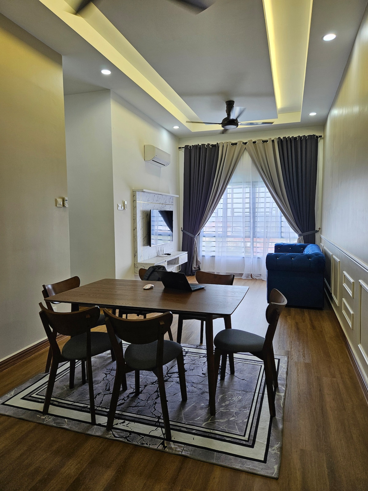 A modern dining area is showcased with a wooden table surrounded by four chairs. Natural light enters through large windows, complemented by elegant curtains. A cozy seating area features a blue sofa, and a television is mounted on the wall opposite the dining setup.