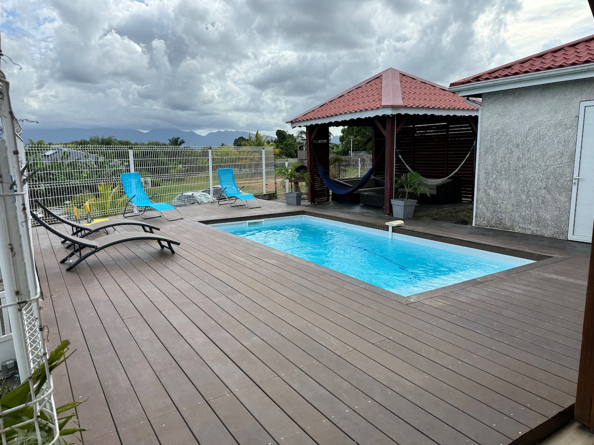 A modern outdoor area features a rectangular swimming pool surrounded by a spacious wooden deck. Two lounge chairs in muted colors are positioned adjacent to the pool, with a shaded gazebo housing a hammock visible in the background. Lush greenery can be seen beyond the fence.
