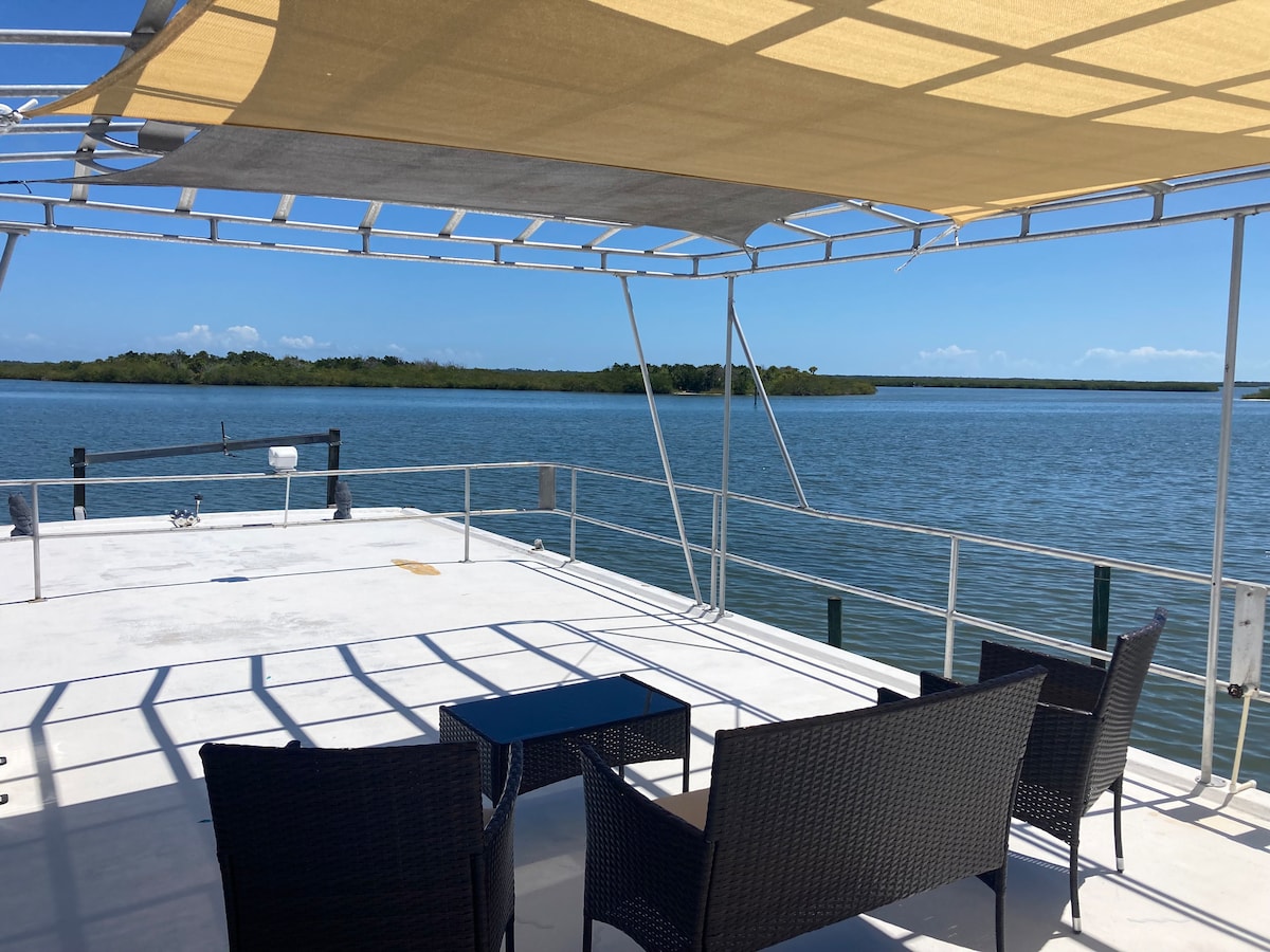 An open deck area is furnished with dark wicker seating and a small table, providing a comfortable space to relax. A large canopy offers shade, while the tranquil waters of the Indian River Lagoon and lush greenery in the distance create a serene waterfront view.