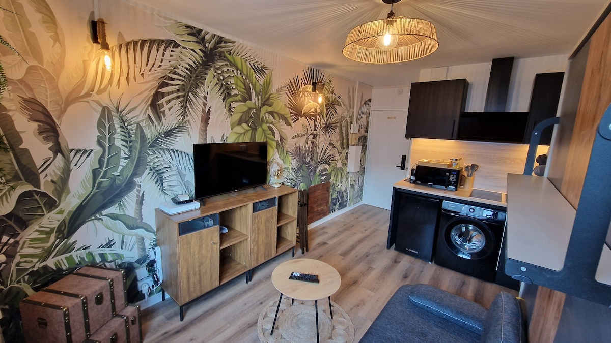 A modern living space is highlighted by tropical-themed wallpaper featuring lush green palm leaves. A cozy seating area is accompanied by a wooden coffee table. A television is mounted on the wall above a stylish cabinet, with appliances including a microwave and washing machine visible in a kitchen area.