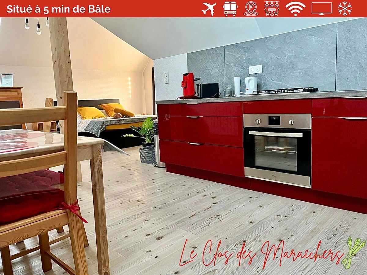 A modern kitchen is featured with red cabinetry and stainless steel appliances, including an oven and stovetop. A wooden dining table with simple chairs is positioned nearby, creating a sense of openness. In the background, a cozy sleeping area is partially visible.