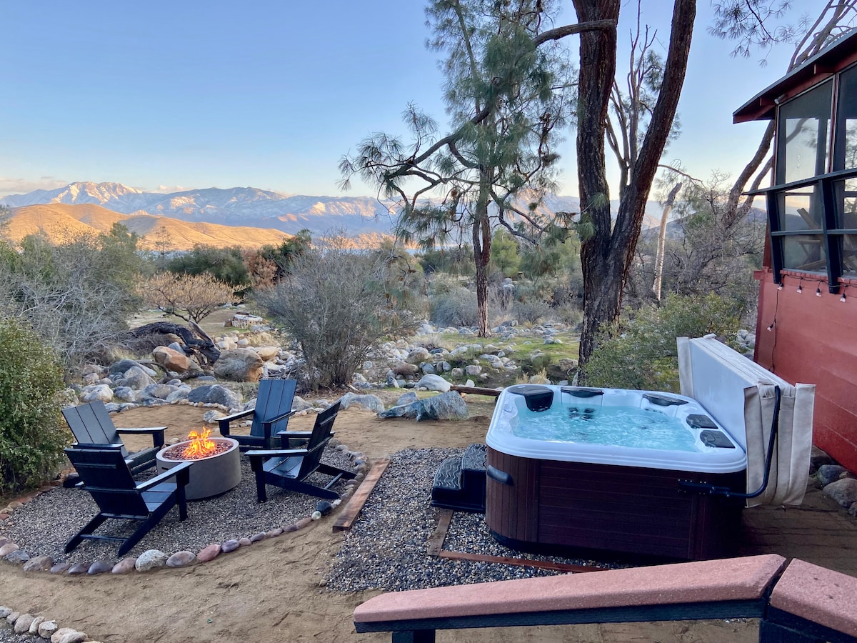 An outdoor area features a hot tub beside a gas fire pit, surrounded by lounge chairs. A serene landscape is visible in the background, showcasing rolling hills under a clear sky with distant mountains.