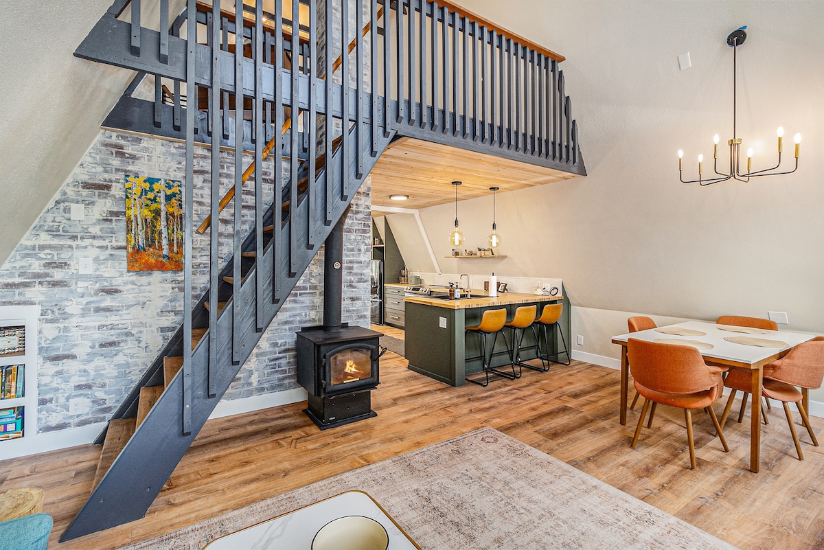 A spacious interior showcases an inviting dining area with orange chairs arranged around a light table. A wooden staircase leads to the loft, with a cozy wood stove providing warmth. Modern kitchen elements are visible in the background, complemented by light wood accents throughout the space.