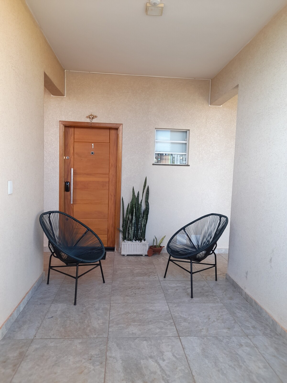 A welcoming entrance area is displayed, featuring a sturdy wooden door and two black chairs positioned on a tiled floor. A tall green plant is situated near the door, contributing to the inviting ambiance of the compact space.