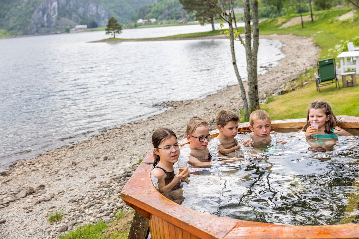 A hot tub sits by the water's edge, surrounded by a natural landscape. Several children are enjoying the water, with trees and a shoreline visible in the background. The serene lake reflects the surrounding greenery, contributing to a peaceful atmosphere.