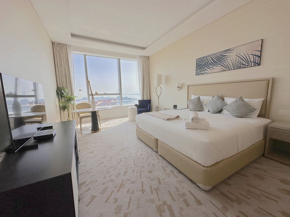 A spacious bedroom features a large bed with decorative pillows and a neutral bedspread. Natural light fills the room through a large window, revealing a view of the outside. A comfortable seating area is present, along with a flat-screen TV and minimalist furnishings.