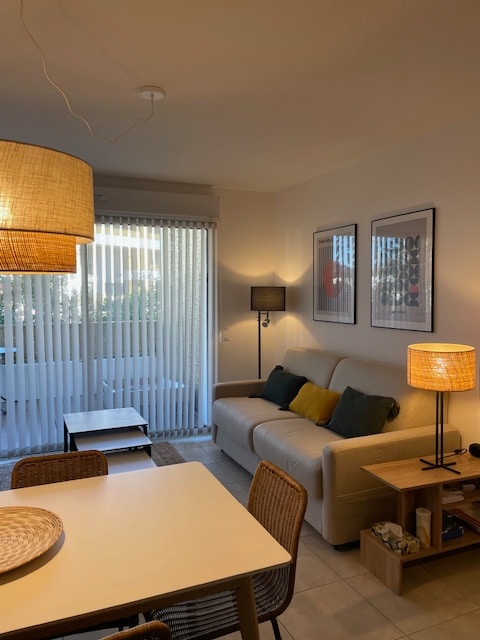 A bright living area features a light-colored sofa adorned with colorful cushions, accompanied by a small coffee table. A dining table with woven chairs is present. Soft lighting from two lamps enhances the inviting atmosphere. Vertical blinds allow natural light to filter in through the glass doors leading to the terrace.
