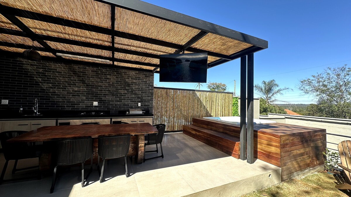 An outdoor area features a shaded dining space with a wooden table and black chairs. A wall-mounted television is visible above an outdoor kitchen equipped with a black tile backsplash. A wooden jacuzzi is positioned nearby, surrounded by a serene atmosphere and clear blue skies.