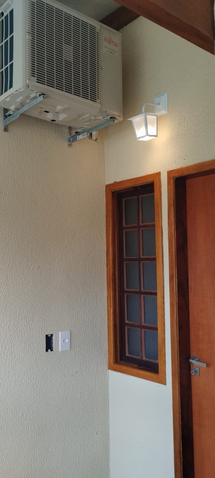 An air conditioning unit is mounted on a textured wall, accompanied by a wall-mounted light fixture. A wooden door with a rectangular window is visible, featuring multiple panes. The overall setting offers a neat and functional entrance space.
