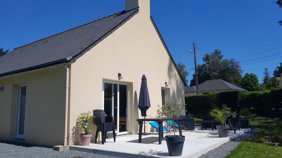 An exterior view of a house is presented, featuring a spacious patio with outdoor furniture including a table, chairs, and an umbrella. Potted plants are arranged around the patio space, and the facade displays a light-colored finish against a clear blue sky.