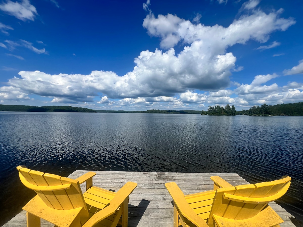 A wooden dock extends into the calm waters of Aylen Lake, flanked by two bright yellow adirondack chairs. The distant shore is visible, framed by trees under a wide sky filled with fluffy clouds, creating a picturesque setting for relaxation.