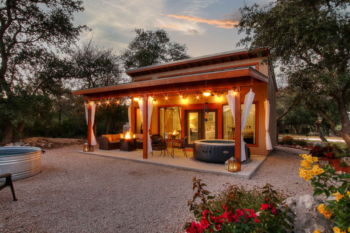 A charming outdoor space is illuminated by string lights, highlighting a gas fire table and a hot tub. Large glass doors lead into the interior. Surrounding trees and flowers add to the serene atmosphere, creating a welcoming environment for relaxation.