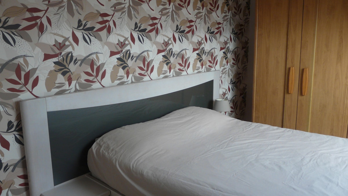 A sleeping area features a spacious bed with a smooth white coverlet against a floral-patterned wall. Natural wood cabinetry provides storage, while a small bedside lamp is positioned next to the bed, enhancing the functional style of the room.