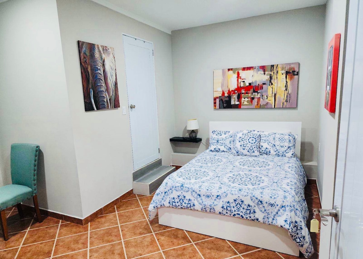 A light-filled bedroom features a full-size bed with a blue and white patterned blanket. Abstract artwork adorns the walls, and a decorative accent chair is positioned nearby. A door leads to an outdoor area, enhancing the space's openness.
