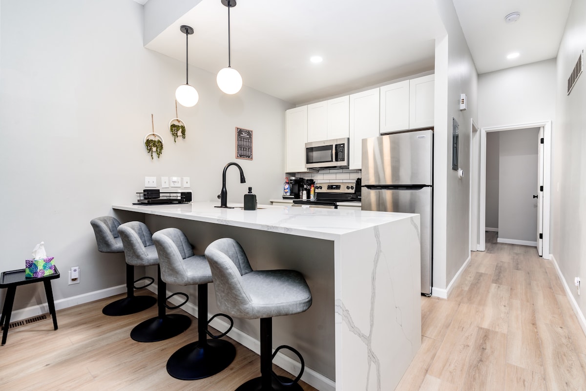 A modern kitchen showcases stainless steel appliances, including a refrigerator and oven. A sleek island countertop with seating for four is paired with stylish bar stools. Warm light illuminates the space, highlighting the clean lines and neutral color palette.