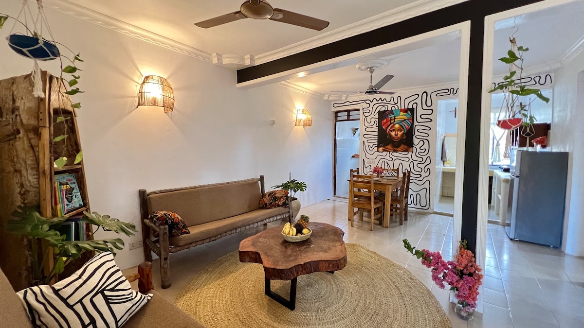 A bright living area features a rustic coffee table and a comfortable sofa adorned with decorative pillows. Plants hang from the ceiling, adding a touch of greenery. A dining table is visible in the background, complemented by modern artwork on the walls.