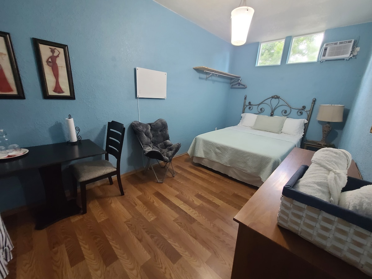 A room features a queen bed with a light blue wall backdrop, complemented by two framed artworks. A small table and chair are arranged near the entrance, while a cozy armchair provides additional seating. Natural light filters in through a small window.