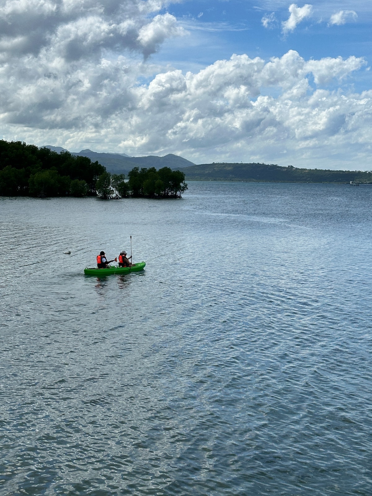 Two individuals are seen kayaking on calm waters, surrounded by lush greenery and distant hills. The sky is filled with fluffy white clouds, reflecting a serene atmosphere over the landscape.