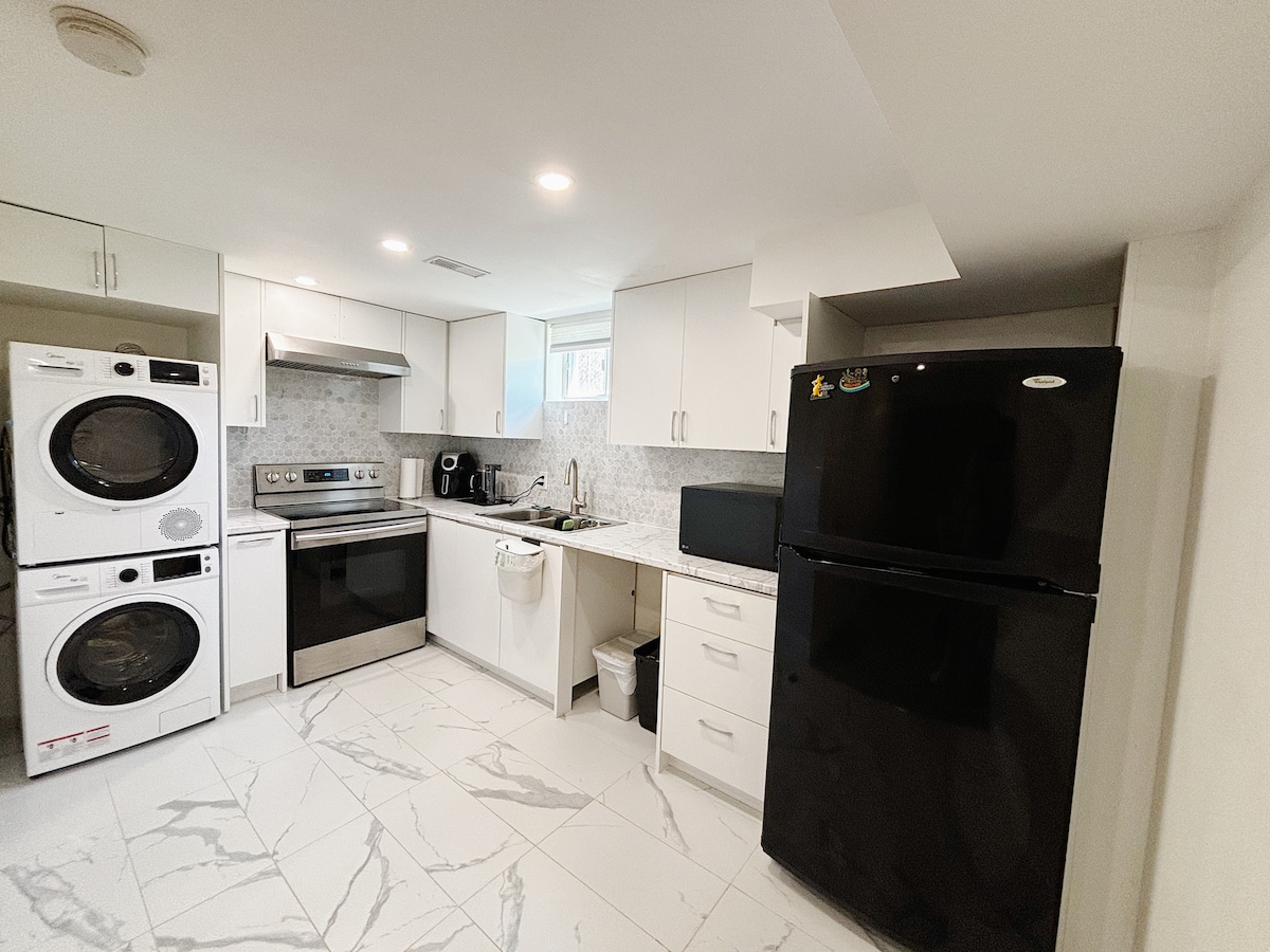 A modern kitchen is visible, featuring stainless steel appliances including an oven, stove, and microwave. A black refrigerator stands against the wall. Laundry machines are positioned to the left, while white cabinetry and marble-patterned flooring complete the space.
