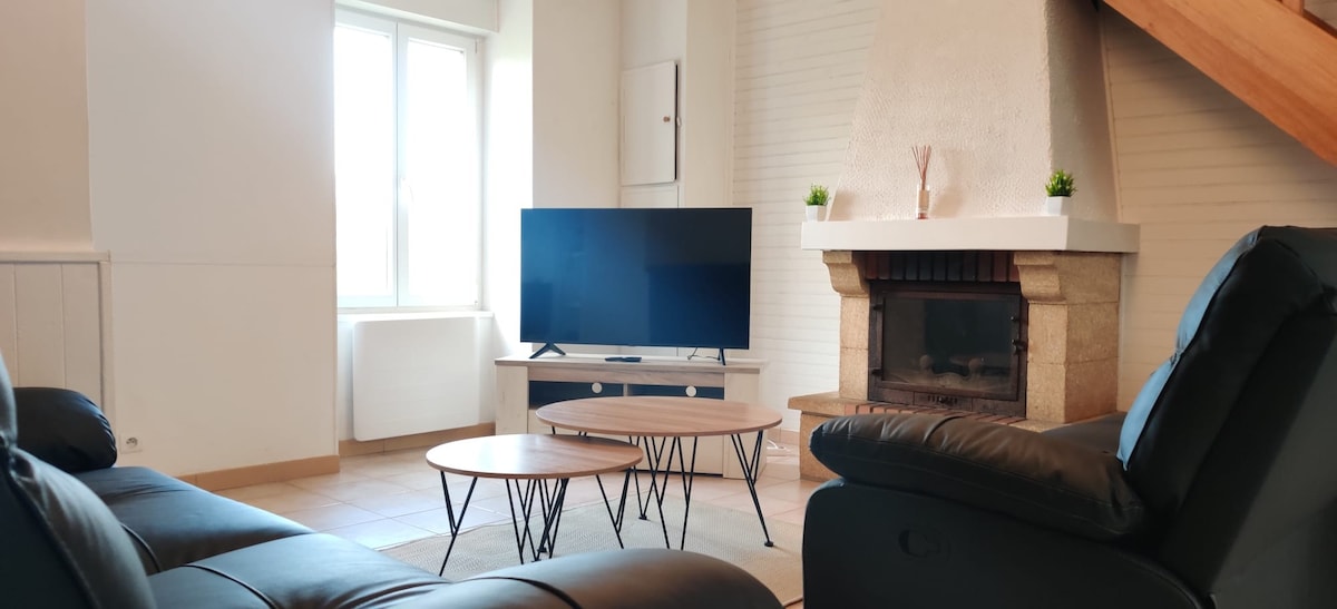 A cozy living room features a large flat-screen television positioned on a low wooden console. Comfortable black leather sofas provide ample seating for relaxation. A simple decorative arrangement sits above the stone fireplace, contributing to the room's welcoming atmosphere. Natural light streams in through the window.
