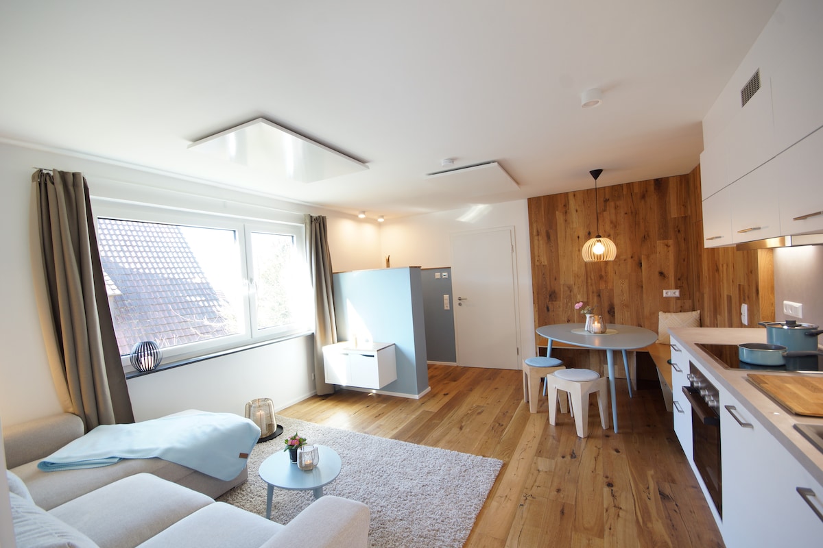 A light-filled living area features a plush couch draped with a soft blanket. A round dining table with chairs is positioned near a window, displaying views of the outdoors. Wooden accents complement modern kitchen cabinetry, creating a warm and inviting space.