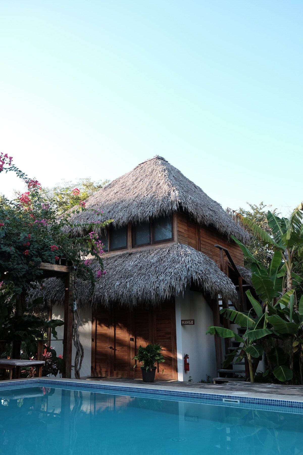 The rustic cabaña features a thatched roof and wooden exterior, surrounded by lush greenery and vibrant flowers. The private pool reflects the clear blue sky, enhancing the serene atmosphere of the property.