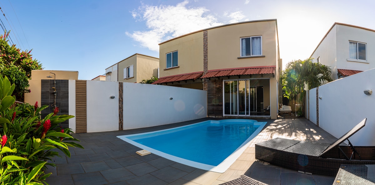 A private outdoor area features a rectangular swimming pool surrounded by paved decking. Lush greenery is visible along the perimeter, and the villa's exterior showcases large glass doors leading into the home. Natural sunlight floods the space, enhancing its serene ambiance.