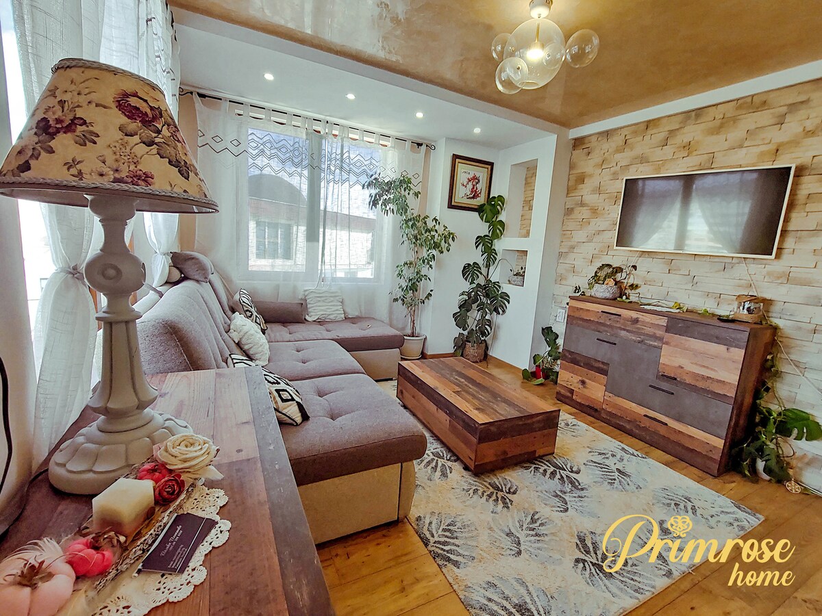 A modern living area features a comfortable sectional sofa, complemented by a wooden coffee table and a TV unit. Natural light filters through large windows adorned with sheer curtains, while decorative plants add a touch of greenery to the inviting space.