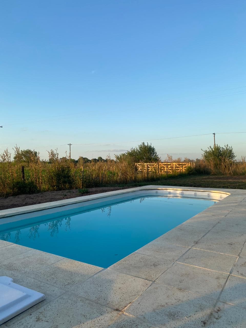 A private pool is framed by a spacious, paved area. Lush greenery surrounds the pool, offering a peaceful view of the open fields beyond. The sky is clear and blue, enhancing the serene atmosphere of the outdoor space.