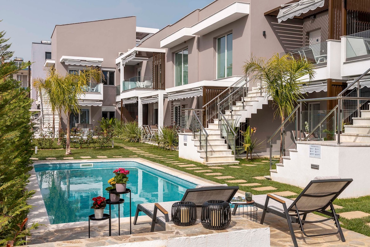 The image showcases a modern apartment complex with multiple units, featuring a central swimming pool surrounded by greenery. Lounge chairs are positioned near the pool, and steps lead to upper-level apartments. Colorful plants and landscaping elements enhance the outdoor space.