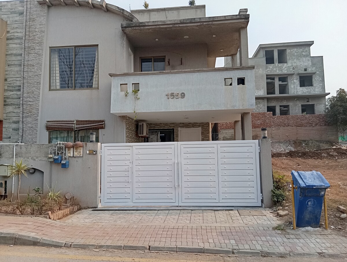 The exterior of the townhouse is displayed, featuring a modern facade with a light grey color scheme. A white gate leads into the property, framed by lush greenery and a paved pathway. In the background, additional construction is visible, indicating ongoing development in the area.