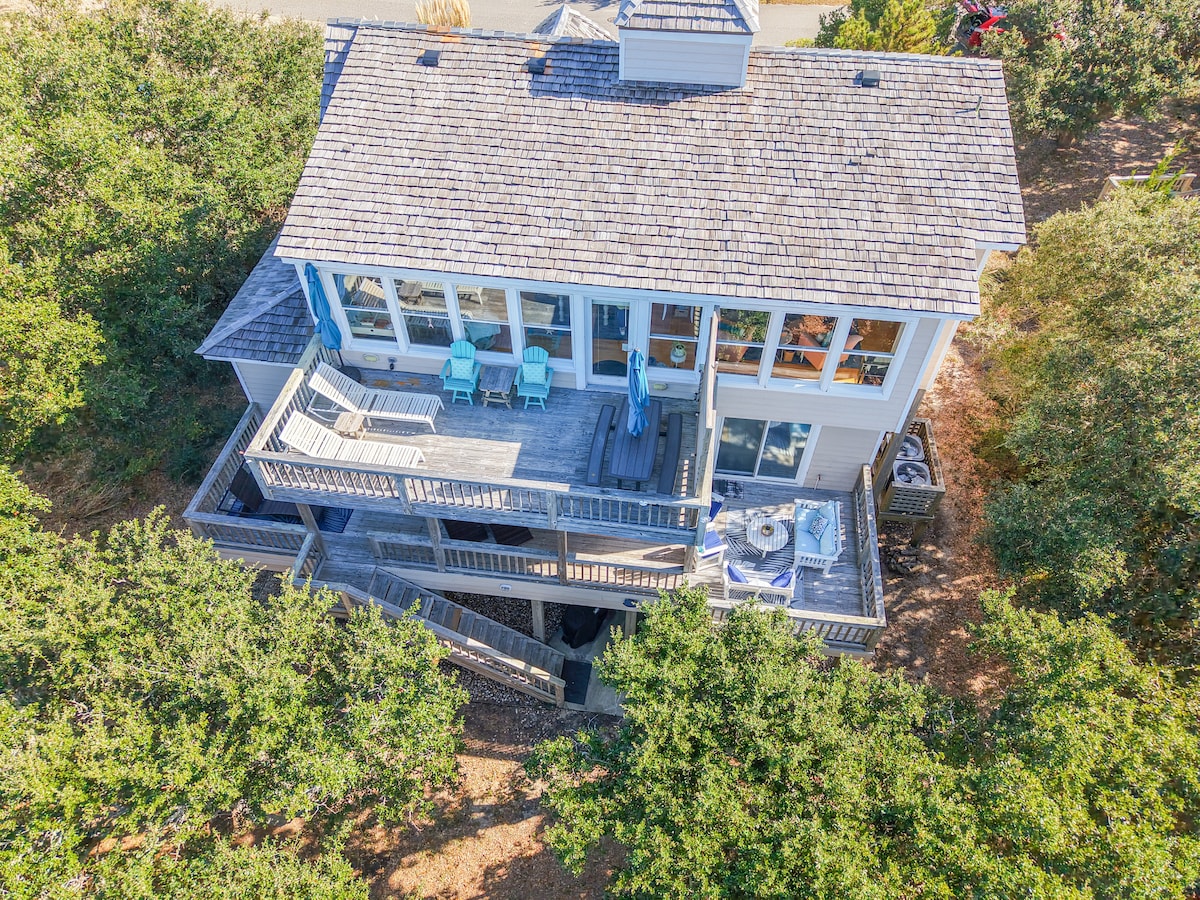 An aerial view of the beachfront home showcases its multi-level deck, featuring lounge chairs and a dining area. Expansive windows allow natural light to fill the interior, while surrounding greenery enhances privacy. The gentle slope towards the beach is also visible, providing a coastal feel.
