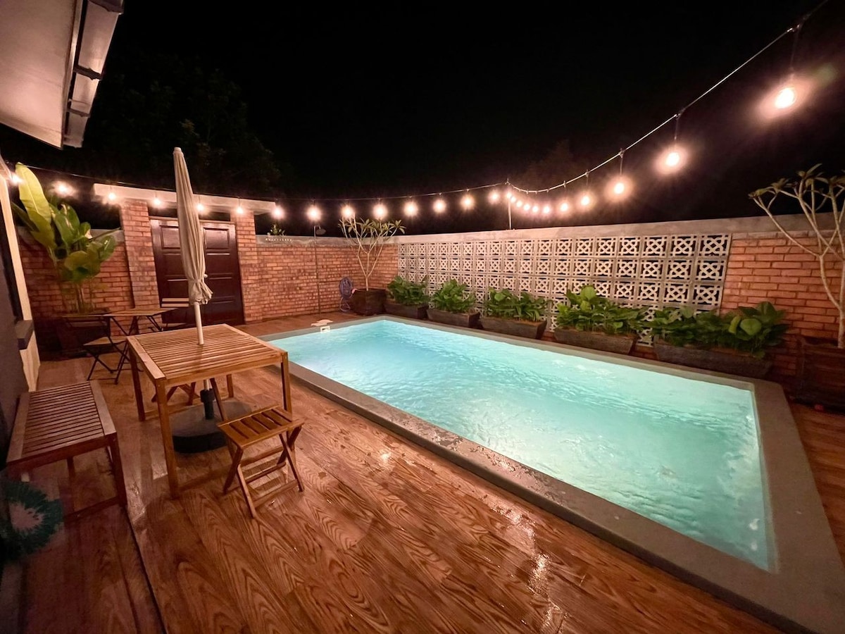 A private swimming pool is surrounded by warm wooden decking and illuminated by string lights. Vibrant greenery lines the edges, creating a serene atmosphere at night. A wooden table and chairs are positioned nearby, inviting relaxation and enjoyment of the outdoor space.