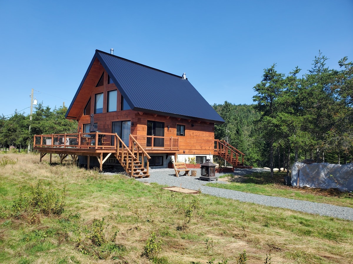 The exterior of a wooden chalet is showcased, featuring a pitched roof and large front deck. Surrounding greenery and a gravel pathway lead to the entrance. The structure is elevated on stilts, offering scenic views of the mountainous landscape.