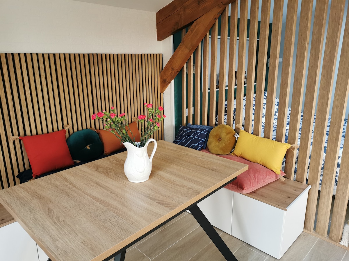 A wooden dining table is centered in the space, highlighted by decorative flowers in a white pitcher. The seating area features a variety of colorful cushions against a backdrop of vertical wooden panels, creating a cozy and inviting nook.
