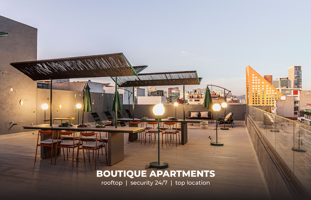 A spacious rooftop terrace is seen, featuring several shaded seating areas with modern furniture. Ambient lighting fixtures are placed thoughtfully around the space. Green umbrellas provide shade, enhancing the comfortable setting while offering city views of the surrounding skyline.