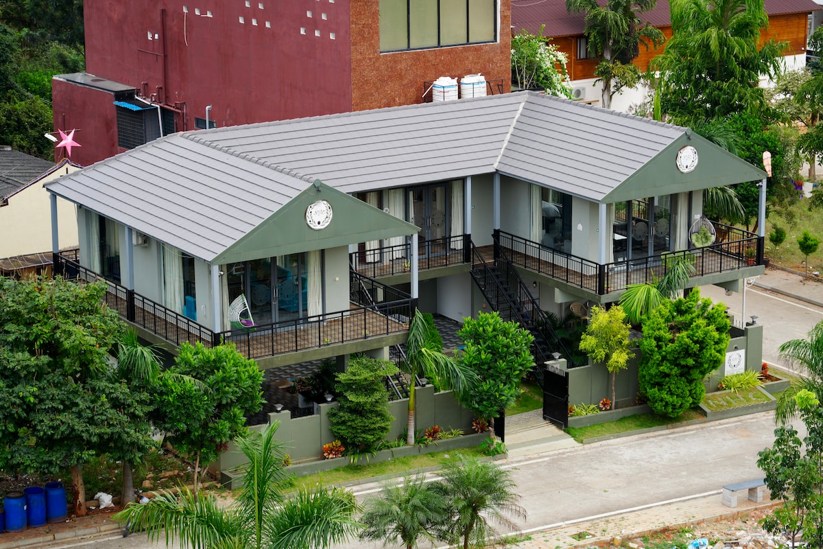 A two-story building features a modern design with a sloped roof and multiple balconies. Lush greenery surrounds the property, complemented by a paved pathway leading to the entrance. The structure is elevated and offers outdoor seating areas, enhancing the inviting ambiance of the cottage.