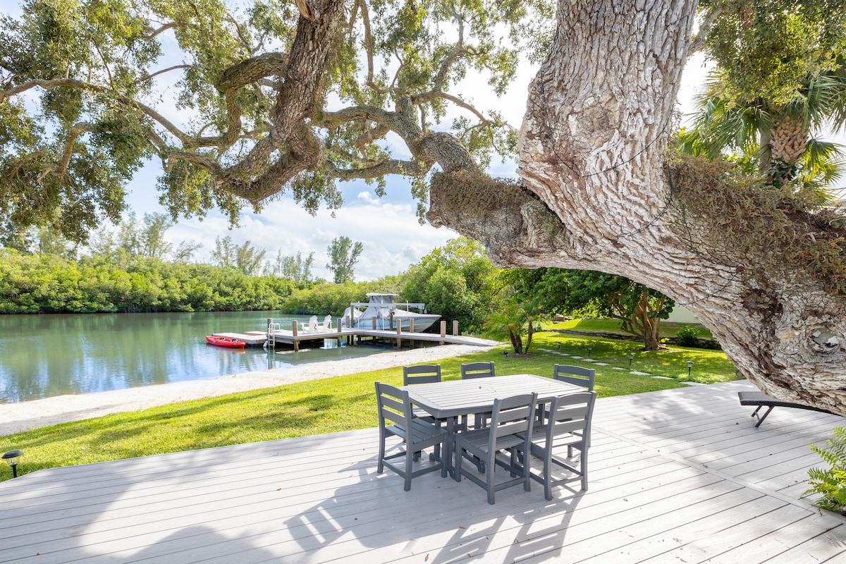 A spacious deck features a wooden dining table surrounded by six chairs, positioned under the shade of a large tree. The serene river views are complemented by a nearby dock and lush greenery, creating a peaceful outdoor setting.