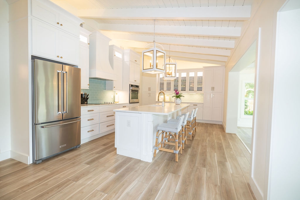 A spacious kitchen is equipped with modern appliances, including a large stainless steel refrigerator. An island with seating is complemented by light-colored cabinetry and elegant light fixtures. Natural light floods the area through windows, enhancing the inviting atmosphere of the open layout.