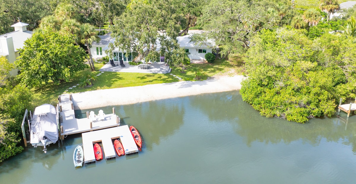 An aerial view showcases a spacious home surrounded by lush greenery, with a sandy area leading to the riverbank. Several colorful kayaks and a boat are docked alongside a wooden platform, emphasizing the property’s direct waterfront access.