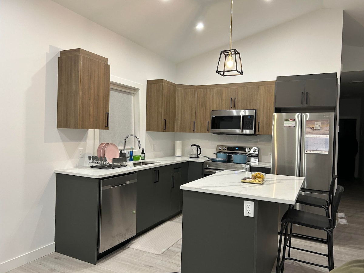 A modern kitchen is presented, featuring dark cabinetry and a light-colored marble countertop. Stainless steel appliances, including a dishwasher and a microwave, are visible. A seated dining area with black stools complements the space, illuminated by a pendant light above.