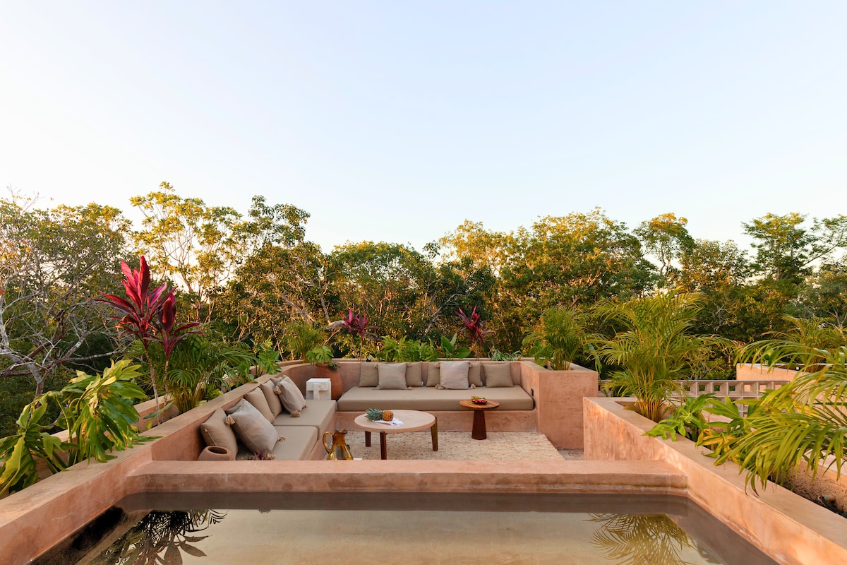 A rooftop lounge area is surrounded by lush tropical foliage, featuring a shallow plunge pool and a circular arrangement of seating. Natural textures and colors create a serene atmosphere, complemented by a small coffee table at the center. The scene captures the tranquility of the jungle setting.