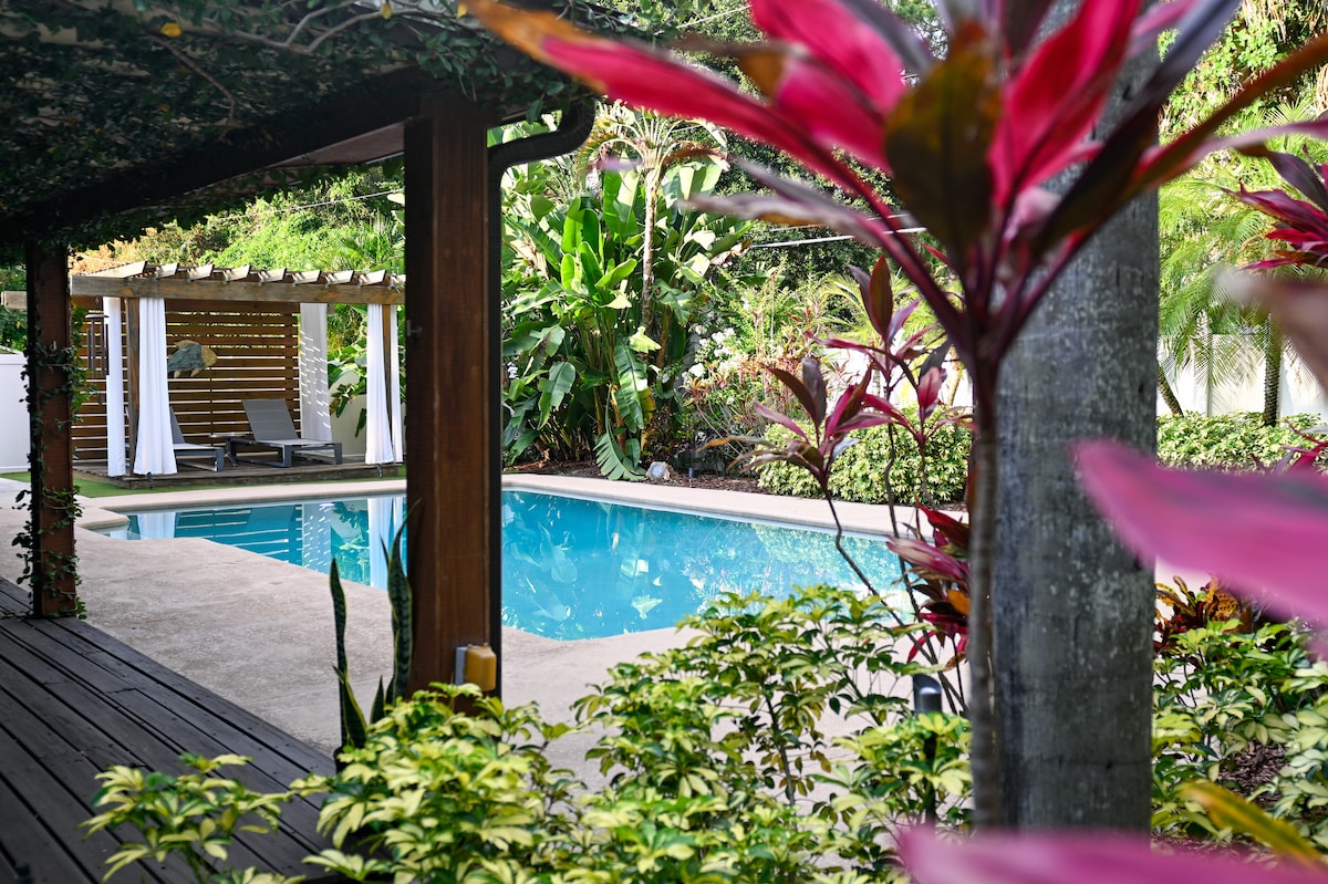 A well-maintained pool area is surrounded by lush greenery and tropical plants. A covered patio offers lounge chairs near the clear water, and poolside privacy is emphasized by vibrant foliage and decorative elements. The serene atmosphere provides a perfect setting for relaxation.