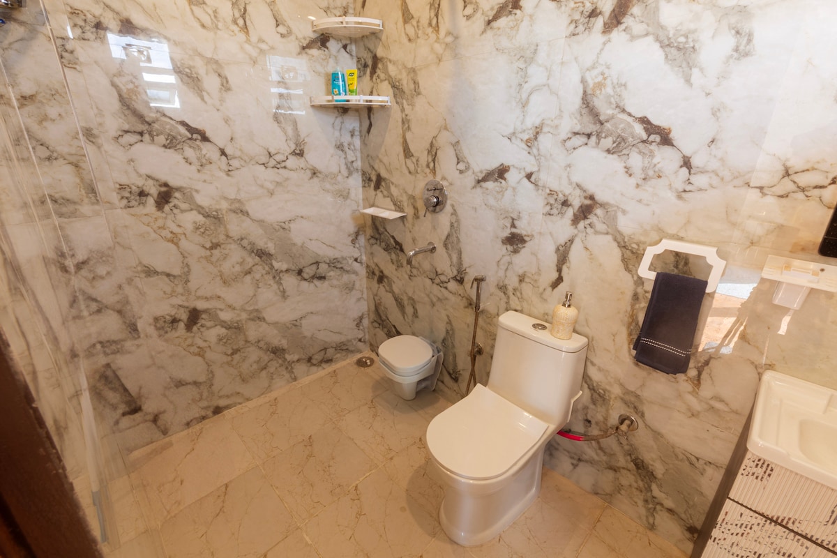 A modern bathroom features elegant marble walls, creating a spacious appearance. It includes a toilet and a separate shower area with glass enclosure. Shelves are mounted on the wall, providing storage for toiletries, while a small sink is positioned nearby.