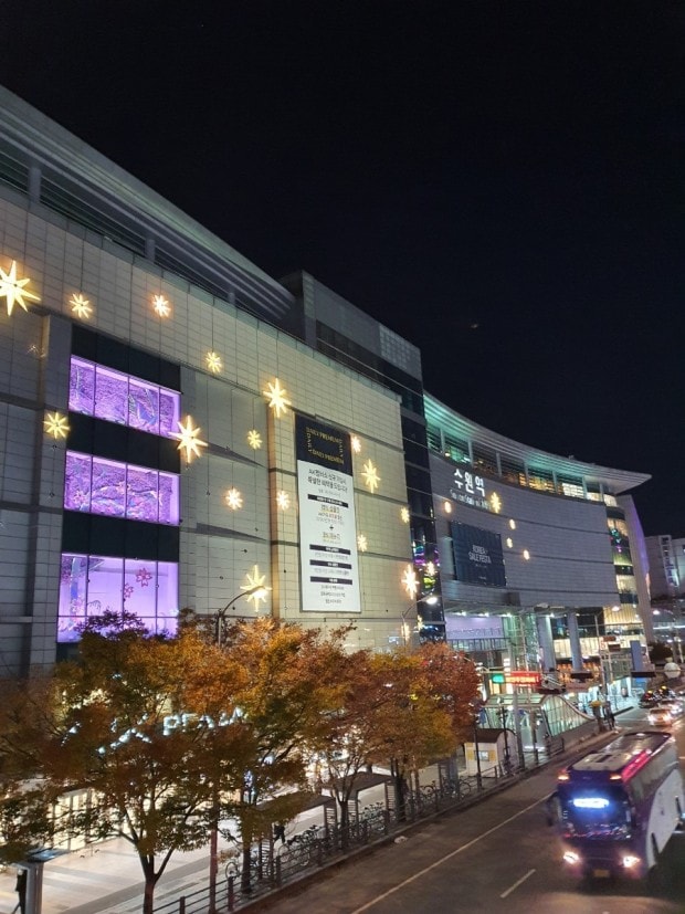 A modern commercial building is illuminated against a night sky, showcasing twinkling star-shaped lights on its façade. Colorful displays are visible through large windows, while trees with autumn leaves line the street, creating a lively atmosphere.