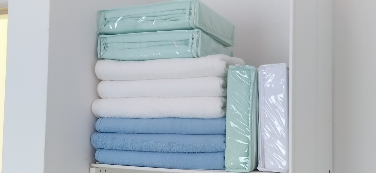 A neatly arranged stack of towels is displayed on a shelf, featuring a mix of pastel colors including white, light blue, and light green. The towels are individually wrapped, indicating cleanliness and readiness for guest use.