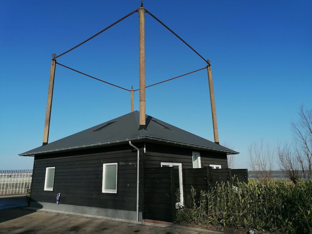 A modern, one-story building features a dark wooden exterior with large windows. The unique architectural design is highlighted by tall, concrete supports positioned above the roof. Surrounding greenery enhances the serene environment, and a clear blue sky provides a bright backdrop.